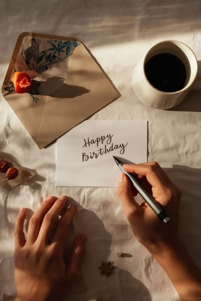 A hand holding a pen to write "Happy Birthday" on a card, with an open envelope and a mug of coffee nearby.